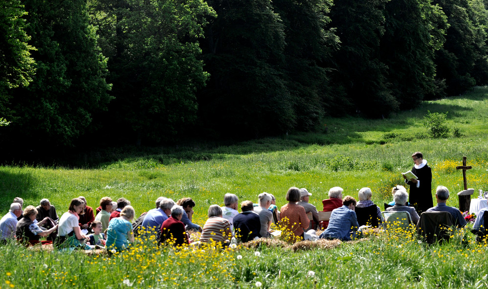 Mennesker sidder på eng i skovkant til friluftsgudstjeneste præst i præstekjole står forrest og læser fra bog